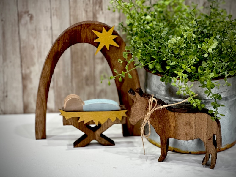 Op de afbeelding: Houten kerststal met een ster, boog en kribbe. Een houten ezel met een touwstrik staat in de buurt. Een metalen emmer met groen gebladerte staat op de achtergrond. De sc&egrave;ne staat op een wit oppervlak.