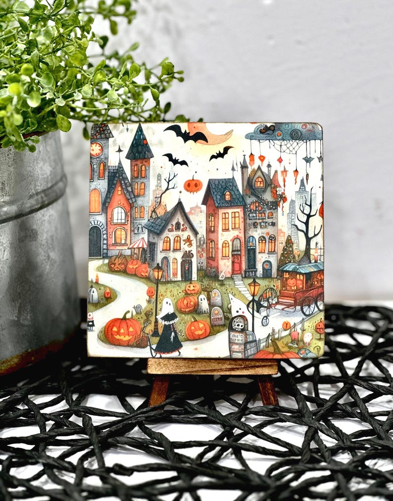 May include: A square Halloween-themed artwork featuring a whimsical town scene with colorful houses, pumpkins, ghosts, and bats. The artwork is displayed on a small wooden easel, perfect for seasonal decor.