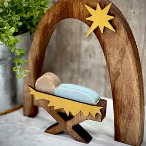 Op de afbeelding: Houten kerststal met een donkerbruine boog en een gouden ster. Binnen de boog is een lichtblauwe wieg met een beige figuur. De wieg wordt ondersteund door een houten standaard met gouden accenten. Een kleine groene plant staat op de achtergrond.