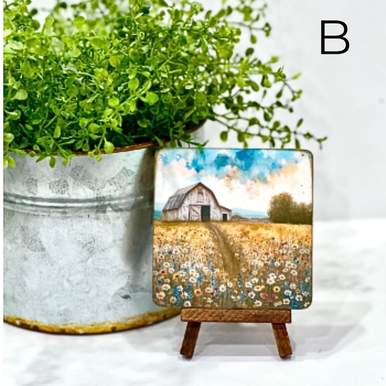 May include: A small painting on a wooden easel depicts a white barn with a red door in a field of wildflowers. The sky is blue with white clouds.