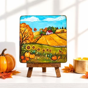 May include: A square painting on a wooden easel depicts a fall landscape with a farmhouse, sunflowers, and pumpkins. The artwork features a blue sky, rolling hills, and a red bird perched on a fence. A pumpkin and candle are in the foreground.