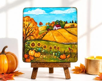 May include: A square painting on a wooden easel depicts an autumnal landscape with a farmhouse, sunflowers, and pumpkins. The artwork features a blue sky, rolling hills, and a red bird. A pumpkin and candle are in the foreground.
