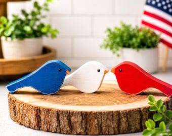 May include: Three wooden bird figurines in patriotic colors: blue, white, and red. The birds are perched on a round wooden slice, with greenery and an American flag in the background. Each bird has a small orange beak and black eye.