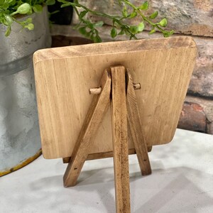 May include: A small wooden easel with a rectangular wooden sign. The easel is made of natural wood with a light brown finish. The sign is blank and ready to be decorated.