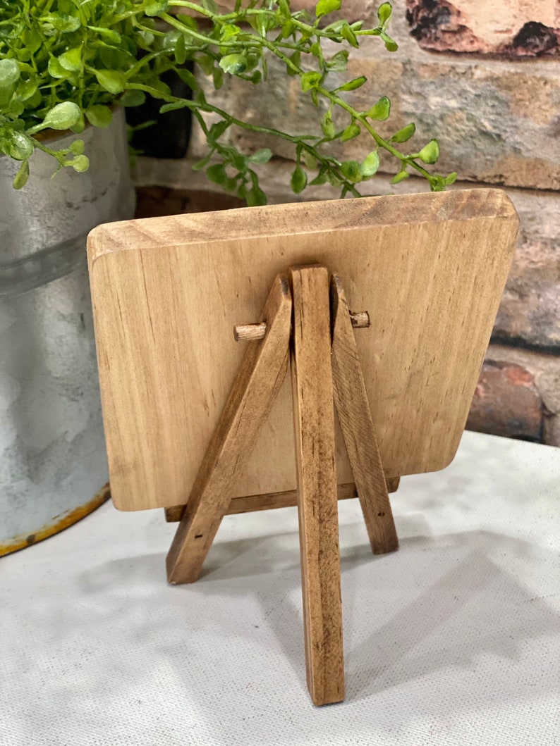 May include: A small wooden easel with a rectangular wooden sign. The easel is made of natural wood with a light brown finish. The sign is blank and ready to be decorated.