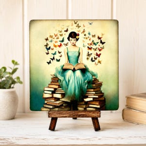 May include: A square art print on a wooden easel depicts a woman in a teal dress reading a book atop a stack of books. Butterflies of various colors surround her. The background is a gradient of green and yellow.