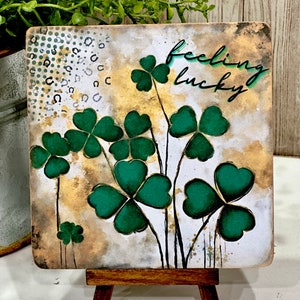 May include: A square canvas with a watercolor background featuring green four-leaf clovers and the text "feeling lucky". The canvas is displayed on a small wooden easel.