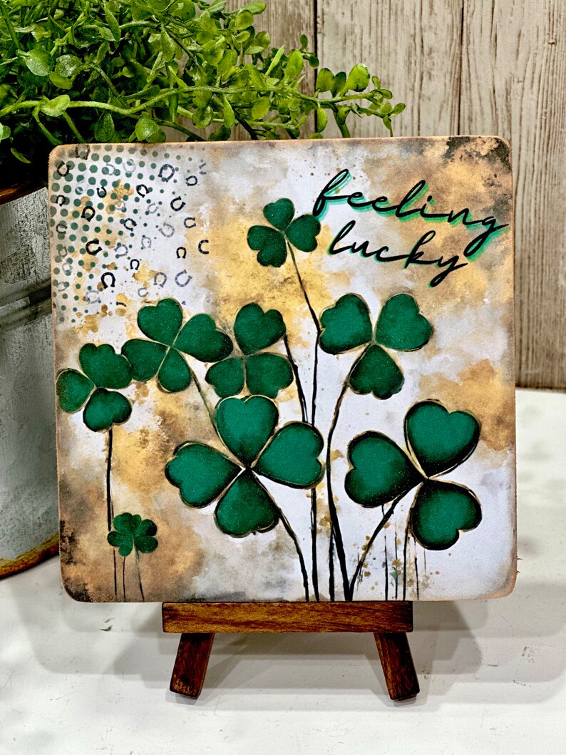 May include: A square canvas with a watercolor background featuring green four-leaf clovers and the text "feeling lucky". The canvas is displayed on a small wooden easel.