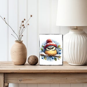 May include: A bluebird wearing a red and white knit hat sits on a branch with blue berries. The bird has a grumpy expression and is surrounded by snow.