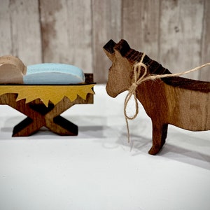 Op de afbeelding: Houten kerststal met een baby in een kribbe en een ezel. De kribbe heeft een geel en bruin ontwerp en de baby ligt op een blauw kussen. De ezel is bruin en bruin met een touwstrik.