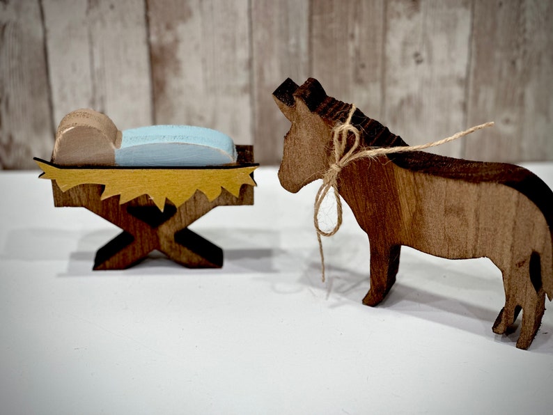 Op de afbeelding: Houten kerststal met een baby in een kribbe en een ezel. De kribbe heeft een geel en bruin ontwerp en de baby ligt op een blauw kussen. De ezel is bruin en bruin met een touwstrik.
