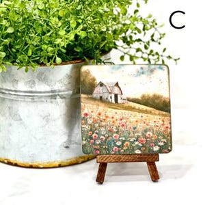 May include: A small wooden easel displaying a square painting of a red barn in a field of wildflowers. The painting has a light blue sky with white clouds.