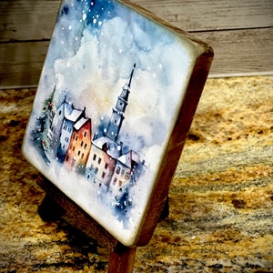 May include: A watercolor painting of a snowy town with a tall church steeple in the center. The painting is on a small wooden easel.