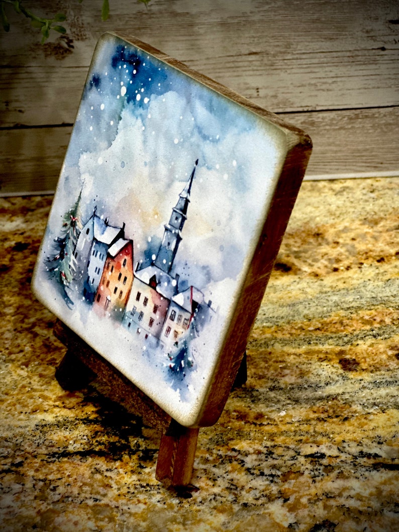 May include: A watercolor painting of a snowy town with a tall church steeple in the center. The painting is on a small wooden easel.
