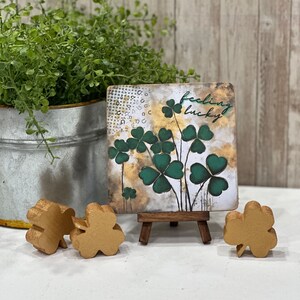 May include: A small wooden sign with a gold background and green shamrocks. The sign says "feeling lucky" and is displayed on a small wooden easel. There are three gold shamrocks on the table.