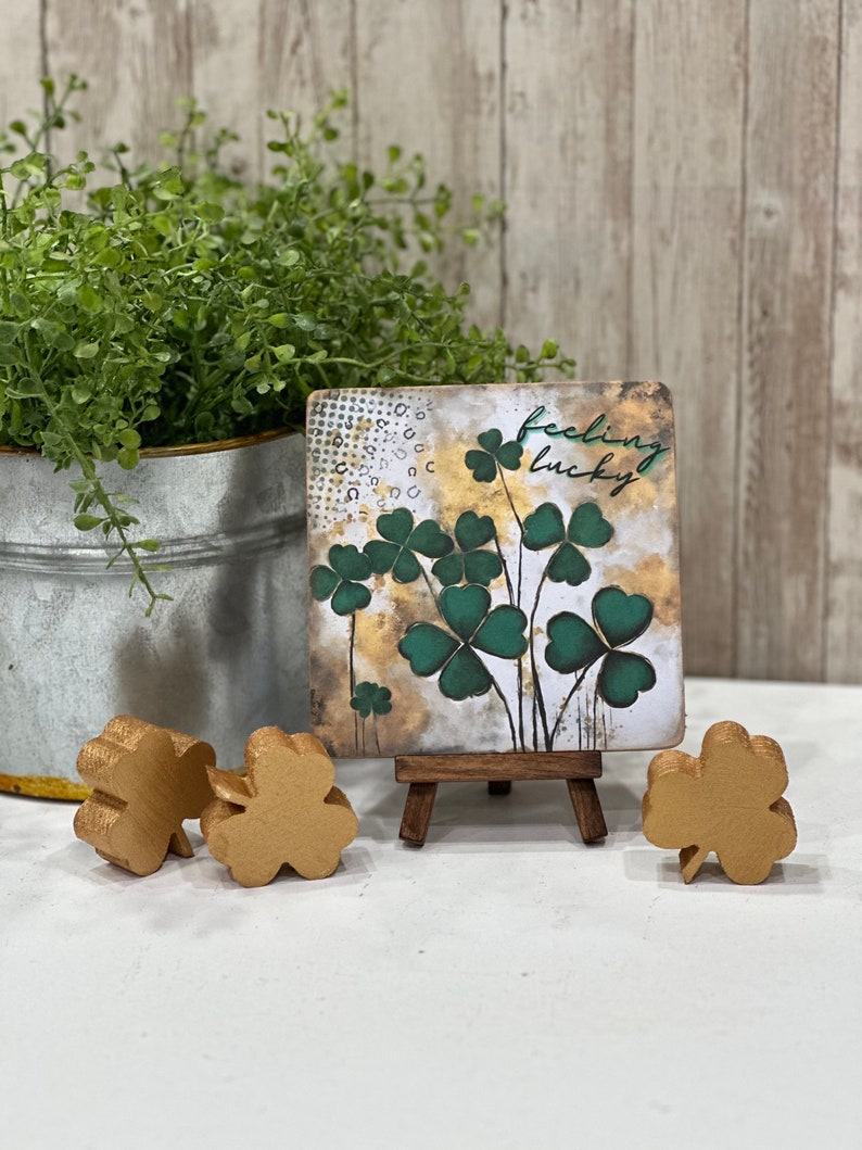 May include: A small wooden sign with a gold background and green shamrocks. The sign says "feeling lucky" and is displayed on a small wooden easel. There are three gold shamrocks on the table.