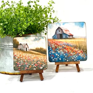 May include: Two small square paintings on wooden easels, each depicting a white barn with a red roof in a field of wildflowers. The painting on the left has a more muted colour palette, while the painting on the right has a brighter, more vibrant colour palette.
