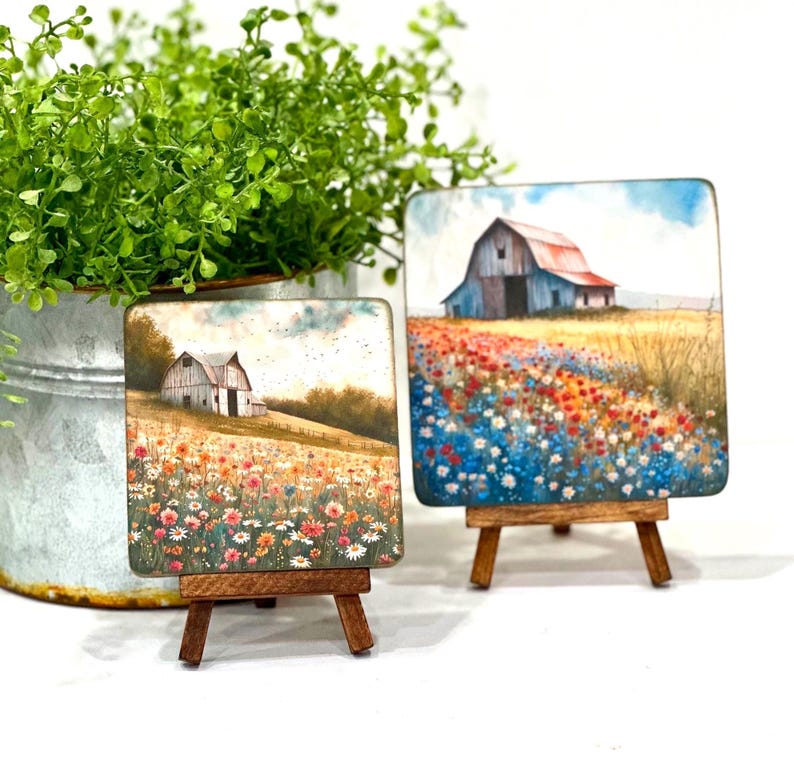 May include: Two small square paintings on wooden easels, each depicting a white barn with a red roof in a field of wildflowers. The painting on the left has a more muted colour palette, while the painting on the right has a brighter, more vibrant colour palette.