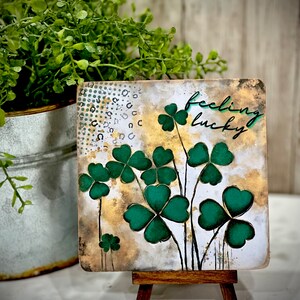 May include: A small wooden easel displaying a square canvas with a watercolor painting of green four-leaf clovers and the text "feeling lucky".