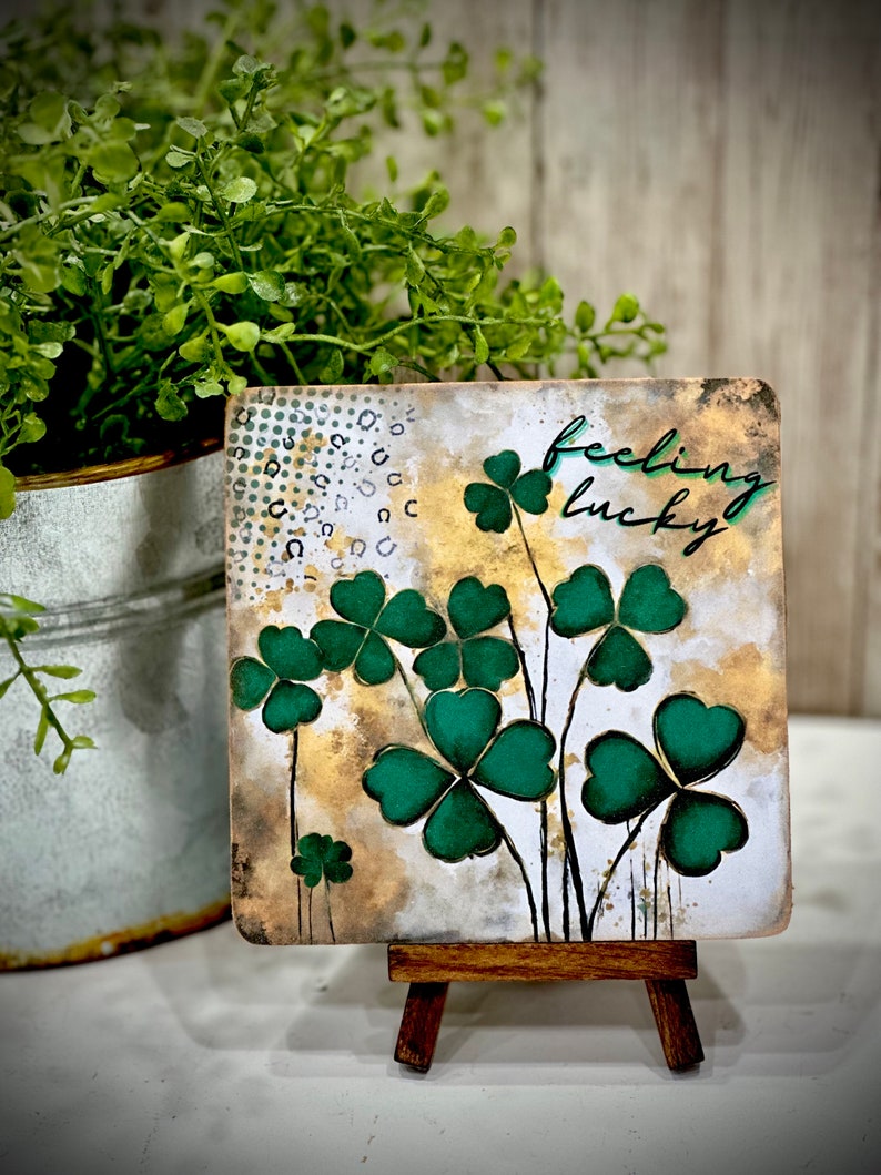 May include: A small wooden easel displaying a square canvas with a watercolor painting of green four-leaf clovers and the text "feeling lucky".