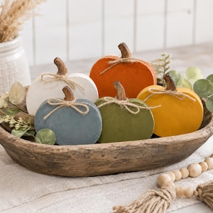 May include: A set of five flat, wooden pumpkin-shaped decorations in white, blue, orange, green, and yellow. Each pumpkin features a brown stem and a tied jute twine bow. The pumpkins are arranged inside a large, oval, dark brown wooden bowl.