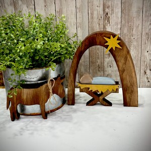 Op de afbeelding: Houten kerststal met een bruine ezel, een kribbe en een boog met een gele ster. De kribbe bevat een babyfiguur met een blauwe deken. De ezel heeft een touw om zijn nek.