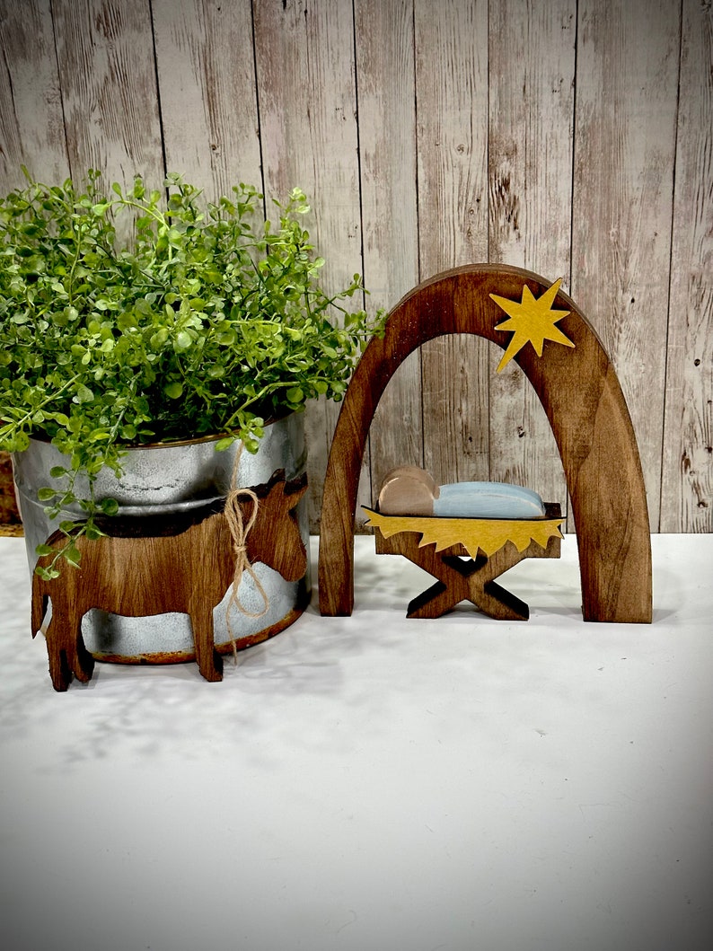 Op de afbeelding: Houten kerststal met een bruine ezel, een kribbe en een boog met een gele ster. De kribbe bevat een babyfiguur met een blauwe deken. De ezel heeft een touw om zijn nek.