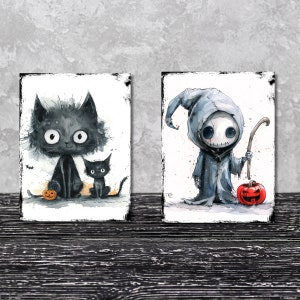 May include: Two rectangular art prints featuring watercolor illustrations. The left print shows two black cats with large eyes and a small orange jack-o'-lantern. The right print depicts a hooded figure in a gray robe holding a staff near a pumpkin.