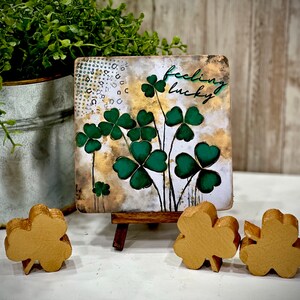 May include: A small wooden sign with a green and gold painted design of shamrocks and the text "feeling lucky". The sign is on a small wooden easel and there are three gold shamrock decorations on a white surface.