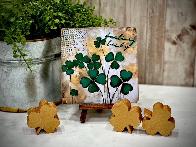 May include: A small wooden sign with a green and gold painted design of shamrocks and the text "feeling lucky". The sign is on a small wooden easel and there are three gold shamrock decorations on a white surface.