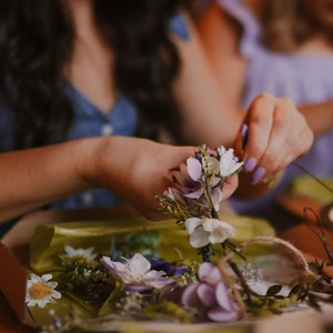 May include: A person is arranging a floral crown with white, purple, and lavender flowers. The flowers are being arranged on a brown twine base. The person is wearing a blue denim top. The background is blurred.