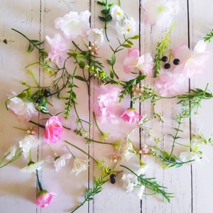 May include: An overhead shot of assorted artificial flowers and greenery. The arrangement includes pink and white blossoms, green leaves, and small black berries, scattered on a white wooden surface.