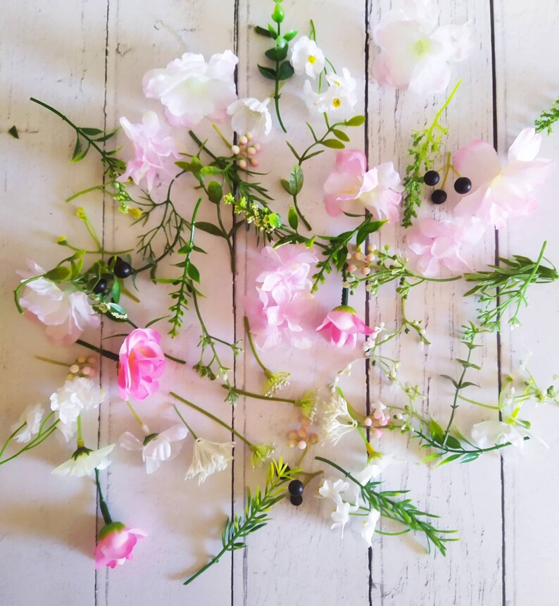 May include: An overhead shot of assorted artificial flowers and greenery. The arrangement includes pink and white blossoms, green leaves, and small black berries, scattered on a white wooden surface.
