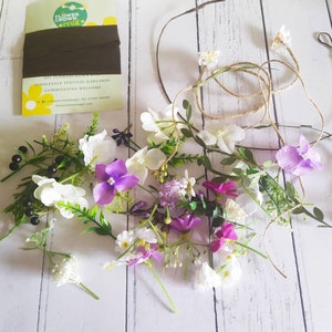 May include: A DIY flower crown kit with various artificial flowers in white, purple, and pink, along with green leaves and a brown ribbon. The kit includes a twine base and a card with the text "Flower Crown Magic".
