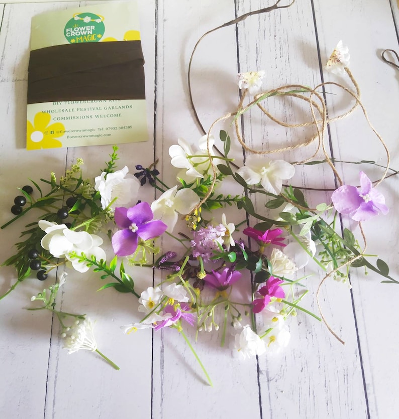 May include: A DIY flower crown kit with various artificial flowers in white, purple, and pink, along with green leaves and a brown ribbon. The kit includes a twine base and a card with the text "Flower Crown Magic".
