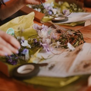 May include: A close-up of a floral crown being assembled. The crown features purple, white, and green flowers. The materials are on a wooden surface with yellow tissue paper and a roll of black tape.
