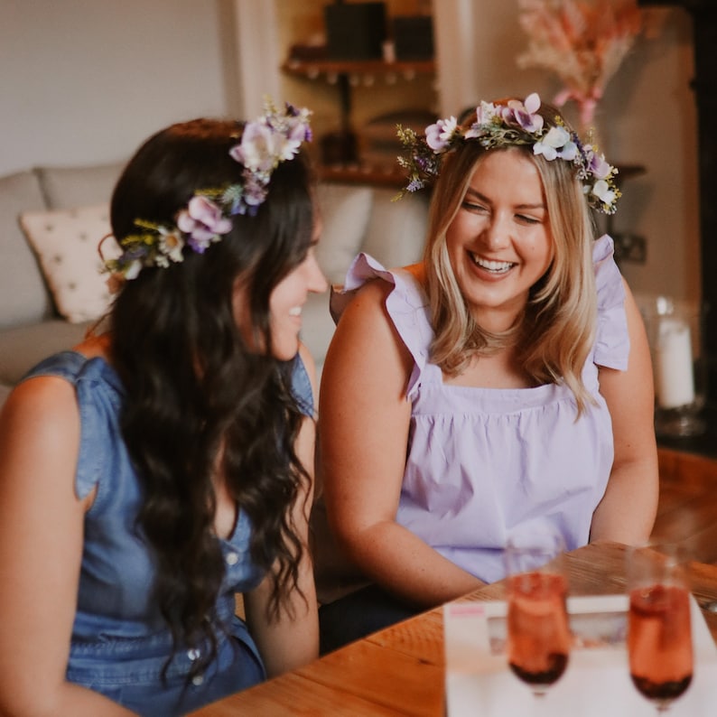 May include: Two people wearing floral crowns and smiling. One person wears a light purple top, the other a blue denim dress. The crowns feature purple and white flowers. Two glasses of a pink beverage sit on a wooden table.