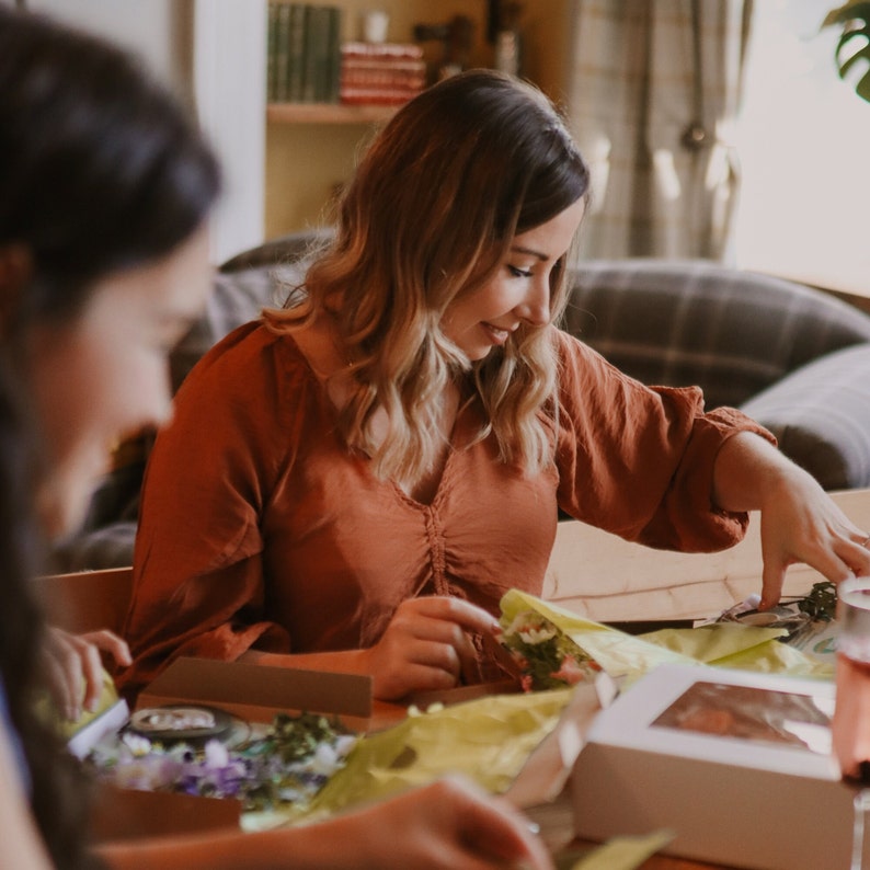 May include: A woman wearing a rust-colored blouse is arranging flowers at a table. The table has various floral arrangements, boxes, and wrapping materials. The scene suggests a craft or floral design activity.
