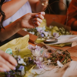 May include: A person is holding a brown wire frame for a flower crown. The table is covered with floral supplies, including purple and white flowers, green leaves, and yellow tissue paper. A roll of black tape is also visible.
