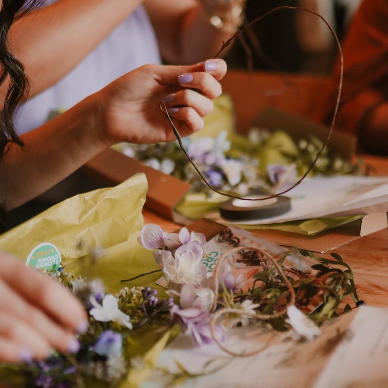 May include: A person is holding a brown wire frame for a flower crown. The table is covered with floral supplies, including purple and white flowers, green leaves, and yellow tissue paper. A roll of black tape is also visible.