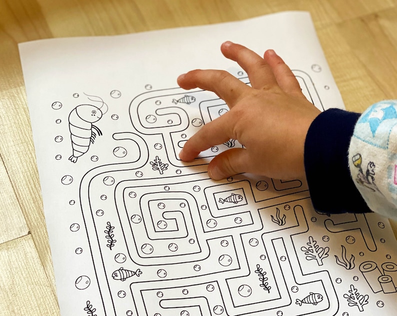 May include: A black and white printable maze activity featuring a shrimp and fish. A child's hand is tracing the maze path.