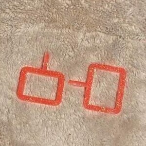 May include: A pair of orange glittery glasses frames. The frames are square-shaped and connected by a small bridge. The glasses are set against a textured, off-white background.