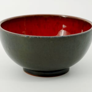 Puede incluir: Un cuenco de cerámica con exterior verde oscuro e interior rojo brillante. El cuenco tiene forma redondeada y se asienta sobre una pequeña base circular. El borde del cuenco es de un tono marrón más oscuro.