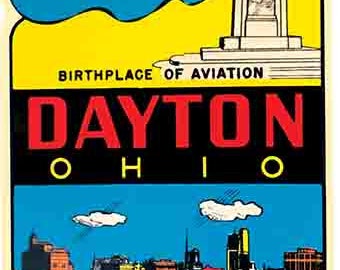 Könnte beinhalten: Ein Vintage-Reisesticker mit gelbem, blauem und schwarzem Design. Der Sticker zeigt ein Doppeldeckerflugzeug, das über eine Stadtlandschaft fliegt. Der Text auf dem Sticker lautet "Birthplace of Aviation Dayton Ohio The City Beautiful".