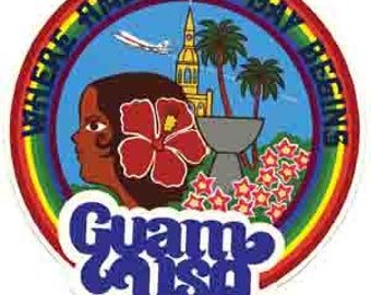 Op de afbeelding: Een ronde, kleurrijke sticker met een regenboogrand. De sticker toont een cartoonillustratie van een vrouwenhoofd met een rode hibiscusbloem in haar haar. De tekst "Where Americans Day Begins" staat in een cirkel rond de afbeelding. De tekst "Guam USA" staat in een groot, gestileerd lettertype onderaan de sticker.