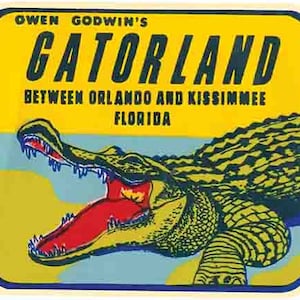 Op de afbeelding: Een geel en blauw vintage-stijl bord met de tekst "Owen Godwin's Gatorland" en "Between Orlando and Kissimmee Florida". Een alligator met een open bek en scherpe tanden is te zien in de onderste helft van het bord.