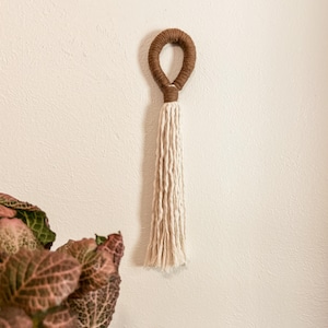 May include: A decorative wall hanging featuring a circular loop wrapped in brown yarn, transitioning into a long, cream-colored fringe made of multiple twisted cotton cords. The piece is suspended vertically, showcasing a contrast between the dark brown top and the light, textured lower section.