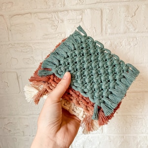 Puede incluir: Un juego de tres posavasos tejidos con flecos. Los posavasos están superpuestos en tonos de verde, naranja y blanco.