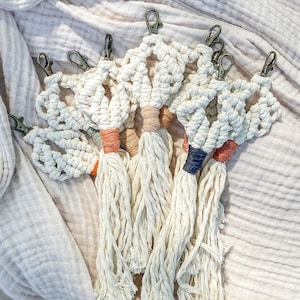 Op de afbeelding: Een set van zes macramé sleutelhangers met kwastjes in verschillende kleuren, waaronder oranje, roze en blauw. De sleutelhangers zijn gemaakt van wit koord en hebben een stervormig ontwerp.