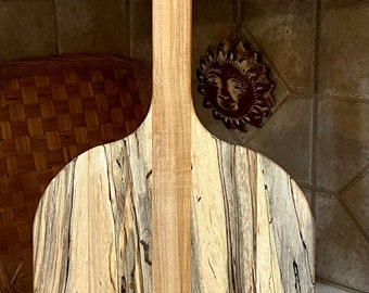 May include: A wooden pizza peel with a light brown handle and a large, oval-shaped paddle made of wood with a blue and brown grain pattern.
